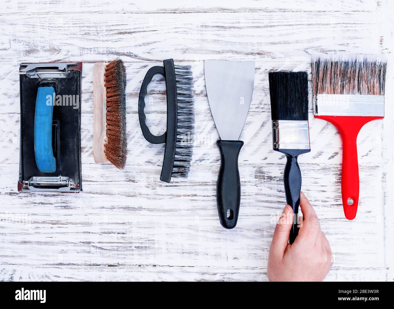 Vecchi attrezzi (pennelli di vernice, spatola, spazzola di filo, supporto di carta di sabbia) e mano che prende uno brus sul bordo bianco. Vista dall'alto, Foto Stock
