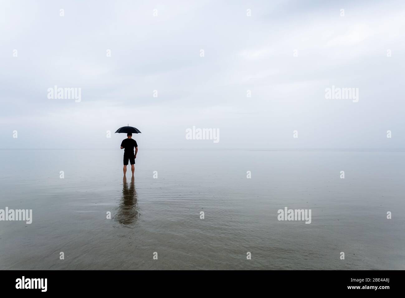 Uomo in piedi su una spiaggia con un ombrello sotto la pioggia. Copia spazio per citazione o testo inspirazione Foto Stock