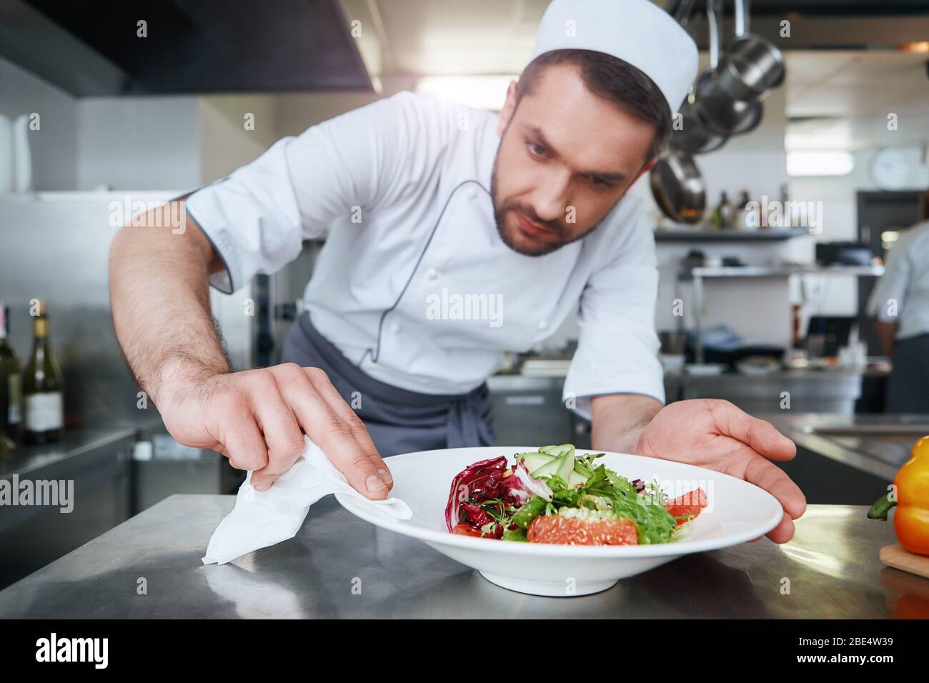 Cuoco caucasico dai capelli scuri che indossa l'insalata di cottura con finiture uniformi bianche. Pulisce i lati del piatto per rendere il piatto più bello Foto Stock Cuoco caucasico dai capelli scuri che indossa l'insalata di cottura con finiture uniformi bianche. Pulisce i lati del piatto per rendere il piatto più bello Foto Stock