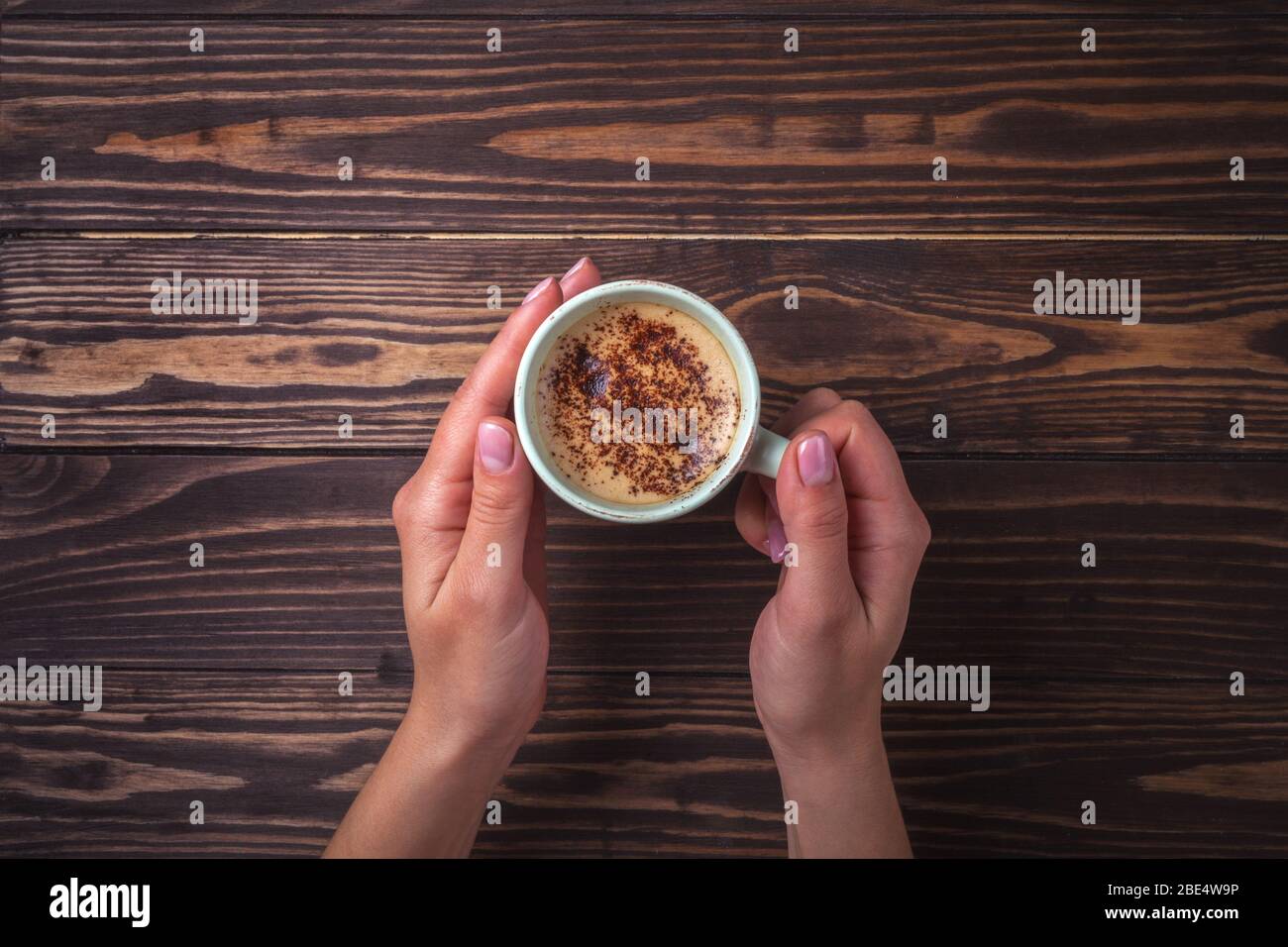 Mani femminili che tengono una tazza di caffè con schiuma su un tavolo di legno, vista dall'alto. Latte o cappuccino con spolverine di cioccolato. Foto Stock