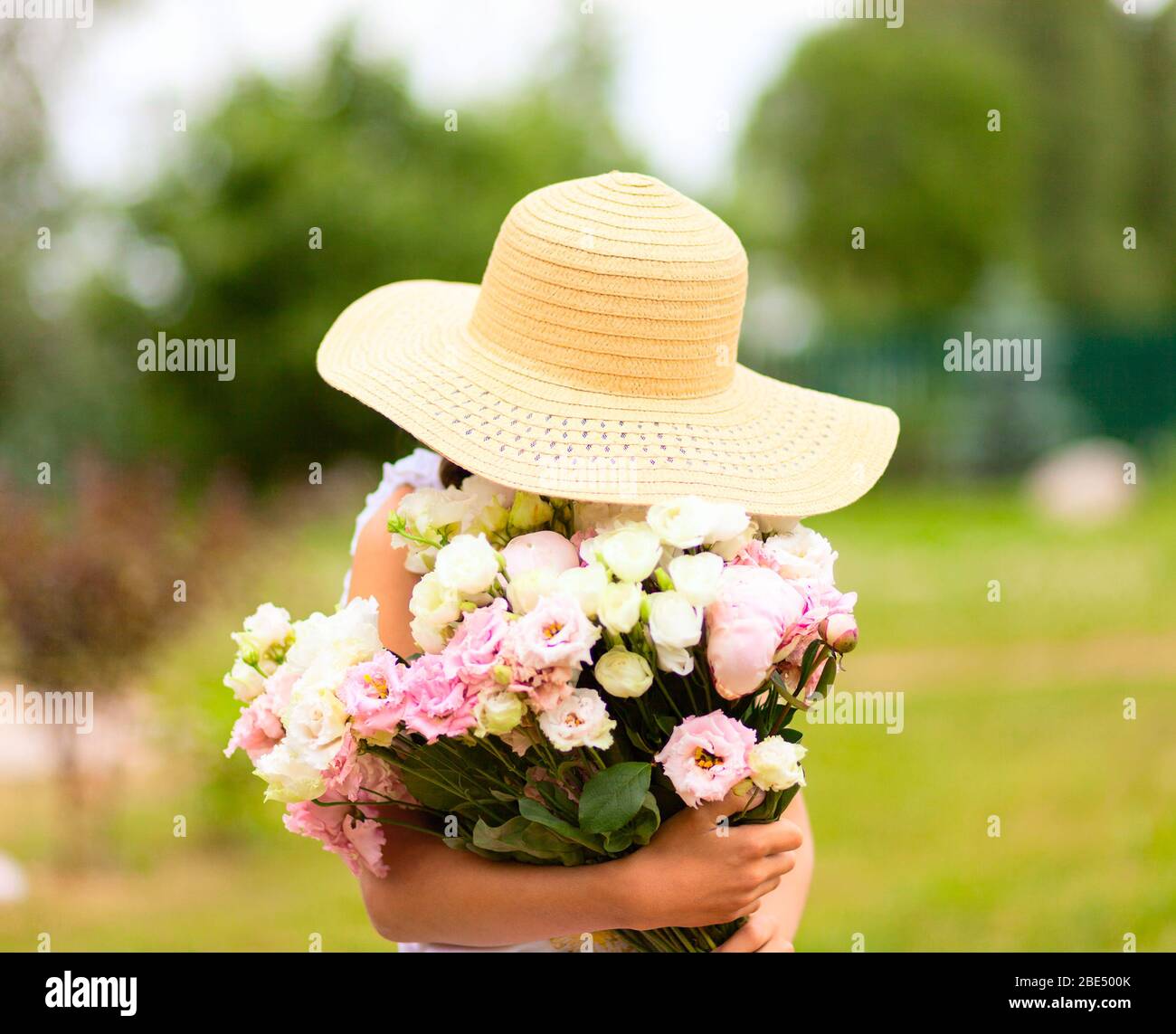 La ragazza tiene un grande bouquet di peonie rosa e bianca davanti a lei. Un bambino in un cappello di paglia con ampio bordo. Gode del profumo dei fiori. Giornata estiva soleggiata. Foto Stock