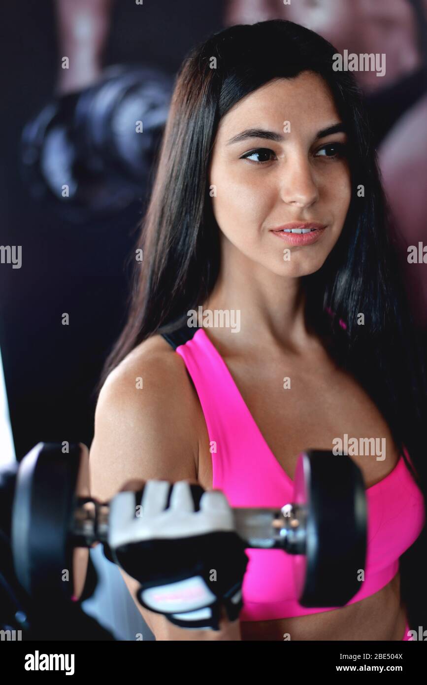 Ragazza sportiva e slim, attrezzature da palestra, bella con un sorriso affascinante, esegue un esercizio con manubri, capelli lunghi, tuta, primo piano Foto Stock