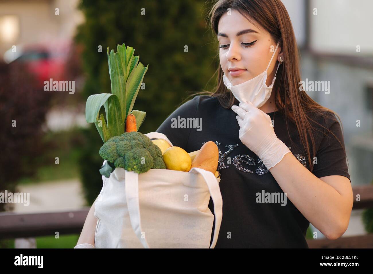 Giovane donna in maschera e guanti tenere sacchetto con cibi freschi, frutta e pane di verdure. Donna dopo la spesa. Proteggersi. Tema coronavirus Foto Stock