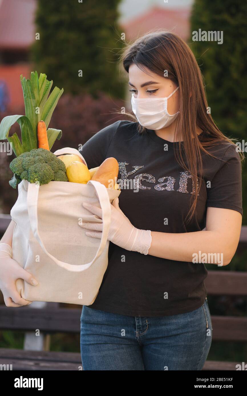 Giovane donna in maschera e guanti tenere sacchetto con cibi freschi, frutta e pane di verdure. Donna dopo la spesa. Proteggersi. Tema coronavirus Foto Stock