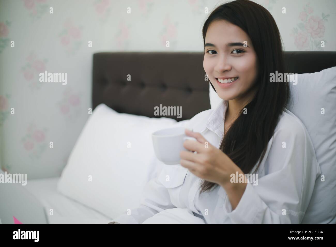 La donna asiatica sta bevendo una tazza di caffè e usando un taccuino per lavorare sul letto.She sorride e ama lavorare a casa. Foto Stock
