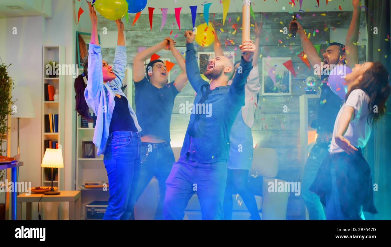 Attraente giovane uomo che lancia i confetti mentre si fa festa con il suo gruppo di amici in una stanza con luci al neon, disco ball e alcol. Scatto al rallentatore Foto Stock