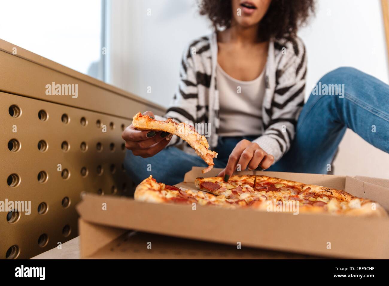 Foto ritagliata di bella giovane donna africana in casa mangiare pizza. Foto Stock