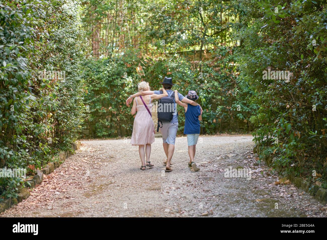 Parte posteriore di piccola famiglia con la madre e due figli che si abbracciano mentre camminano sul sentiero di un parco tra i grandi cespugli verdi Foto Stock