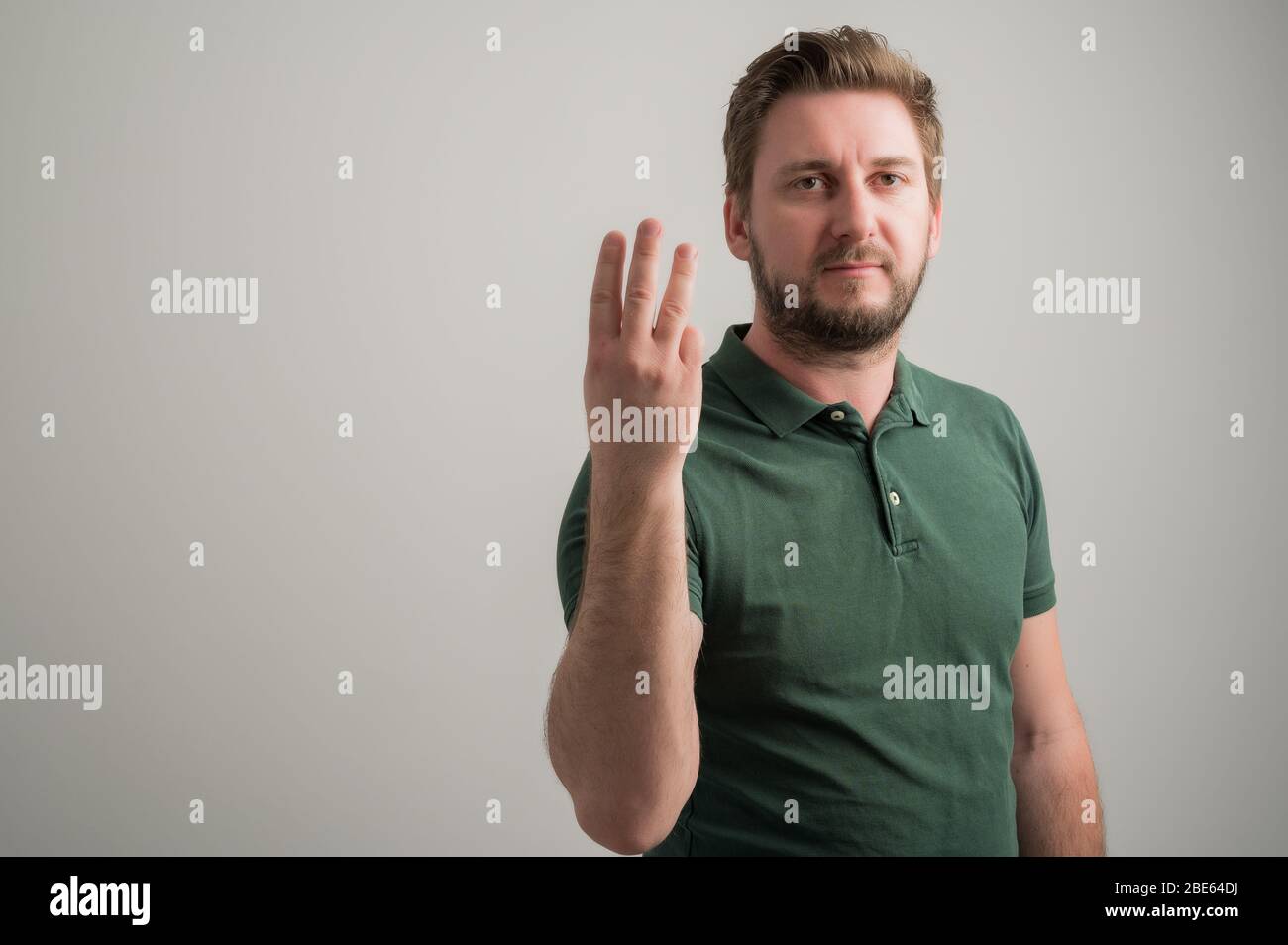 Ritratto di uomo serio elegante attraente con barba spessa, vestito in casual t shirt verde che mostra il numero tre isolato su sfondo grigio con poliziotto Foto Stock