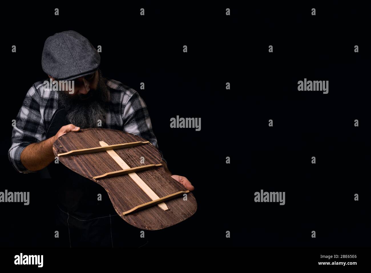 Liutaio guardando il retro della chitarra. Sfondo scuro. Uomo bearded che indossa il cappellino, la camicia a plaid e il grembiule nella sua officina. Foto Stock