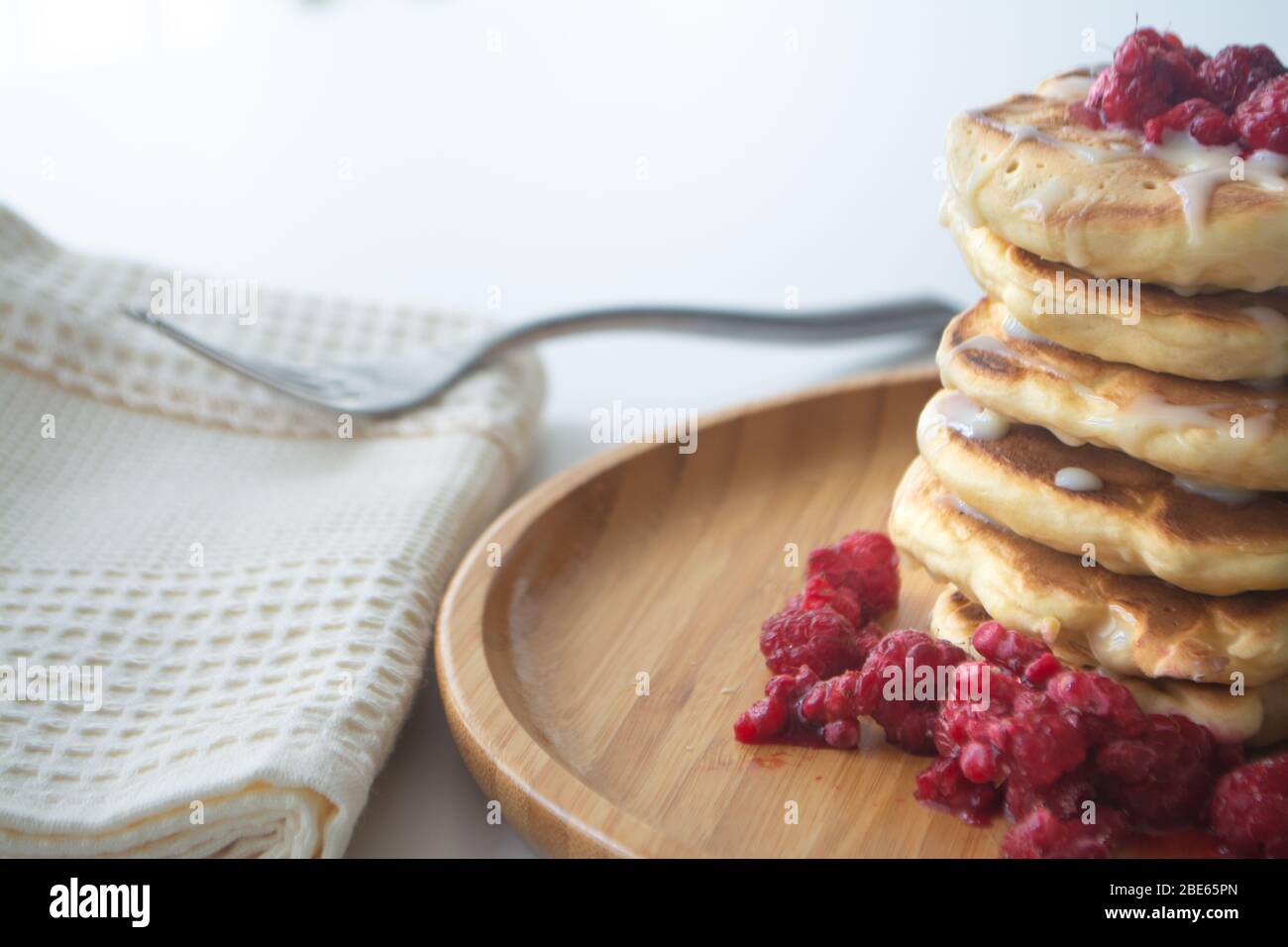 Frittelle soffici con latte condensato e lamponi su un piatto di legno con tovagliolo da cucina e forchetta Foto Stock