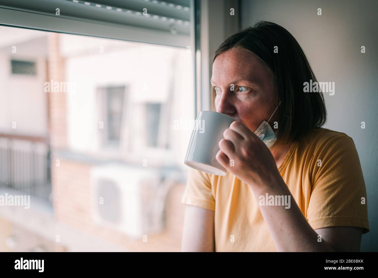 Donna in auto-isolamento durante lo scoppio del virus bere caffè e guardare attraverso la finestra. Persona preoccupata femmina con maschera chirurgica protettiva in soggiorno Foto Stock