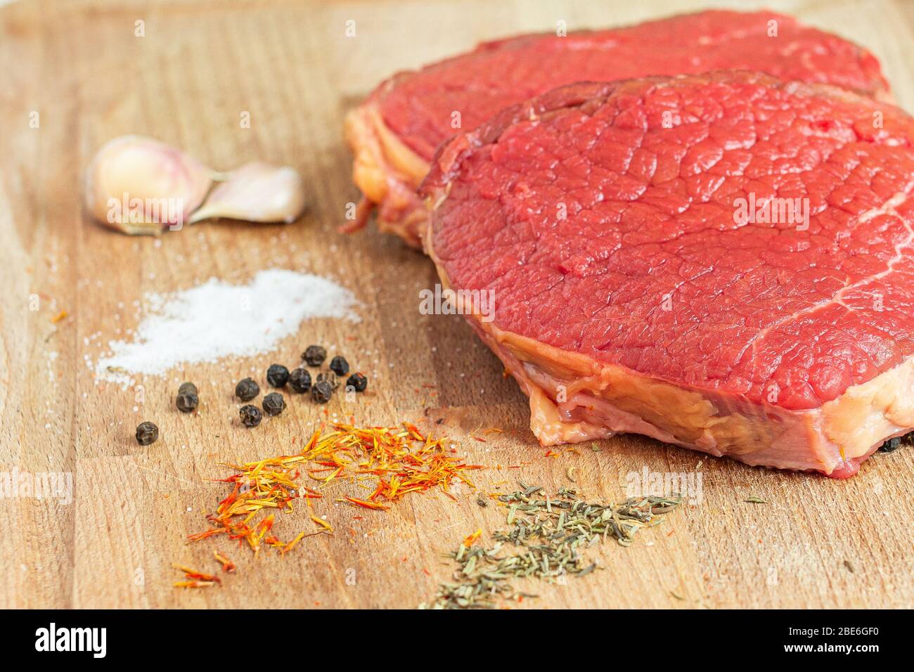 Carne cruda su un tavolo di legno con aglio, sale, pepe nero, zafferano e rosmarino pronti per cucinare. Foto Stock