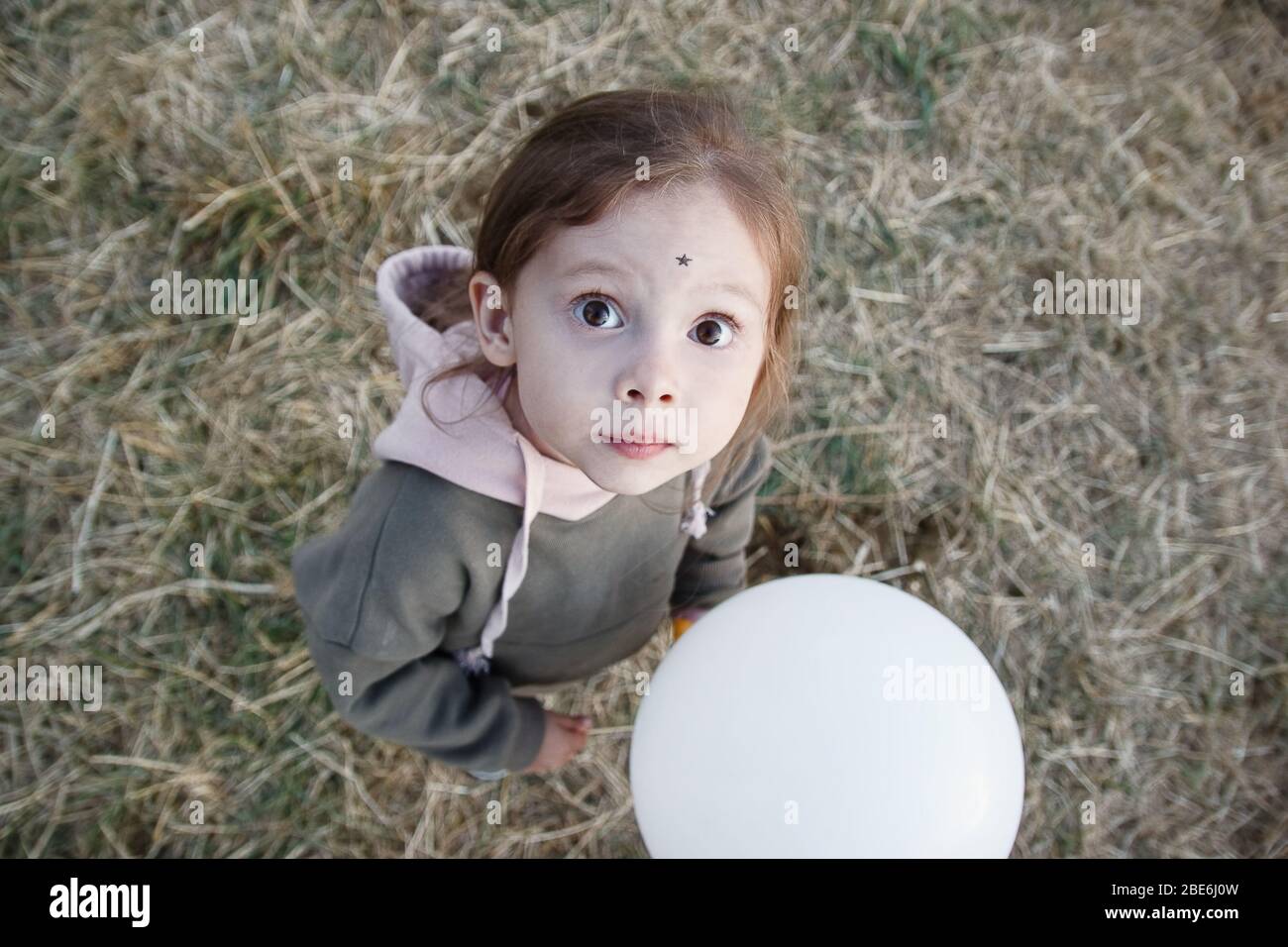 Ritratto carino della bambina con piccola stella nera del corpo arte sul suo viso e baloon bianco in mano. Vista dall'alto, guardando la fotocamera. Attività all'aperto Foto Stock