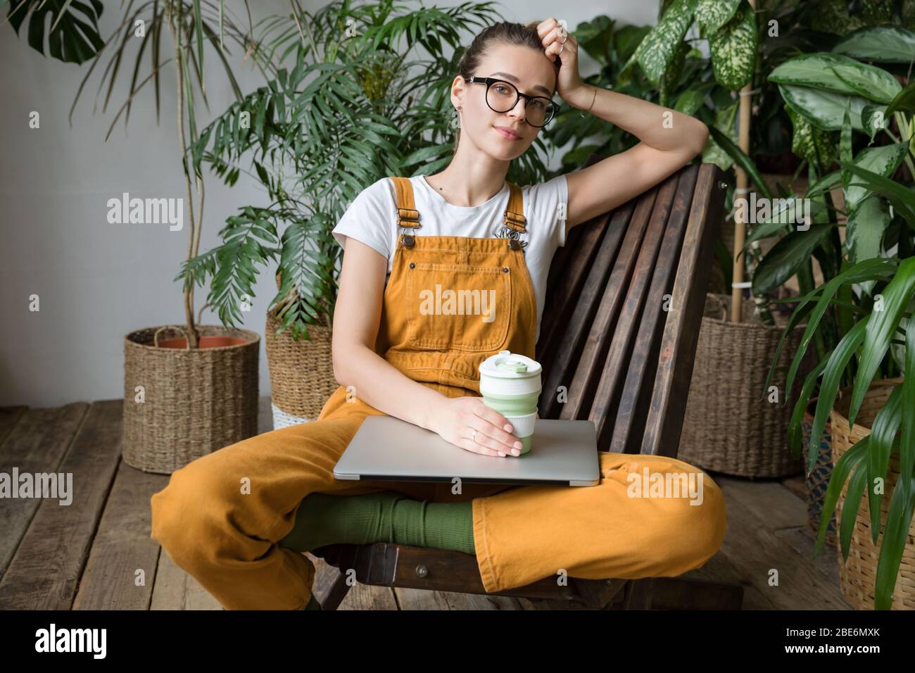 Donna europea giardiniere che indossa tute, riposo dopo il lavoro, seduta su sedia di legno in casa serra, tenere tazza di caffè / tè riutilizzabile, guardando ca Foto Stock