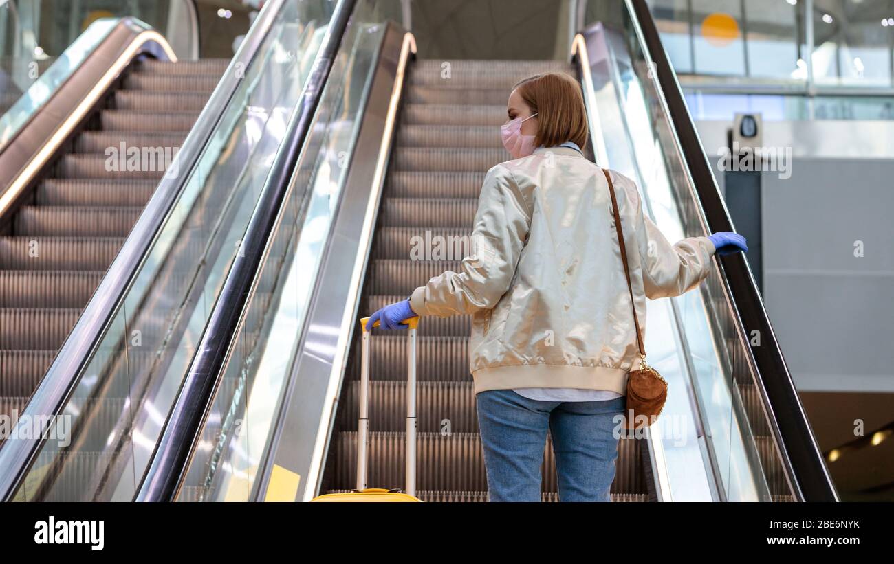 Donna con i bagagli si alza sulla scala mobile, tiene la scala mobile nel terminal dell'aeroporto attraverso guanti in lattice, per proteggersi dal contatto con la ger Foto Stock