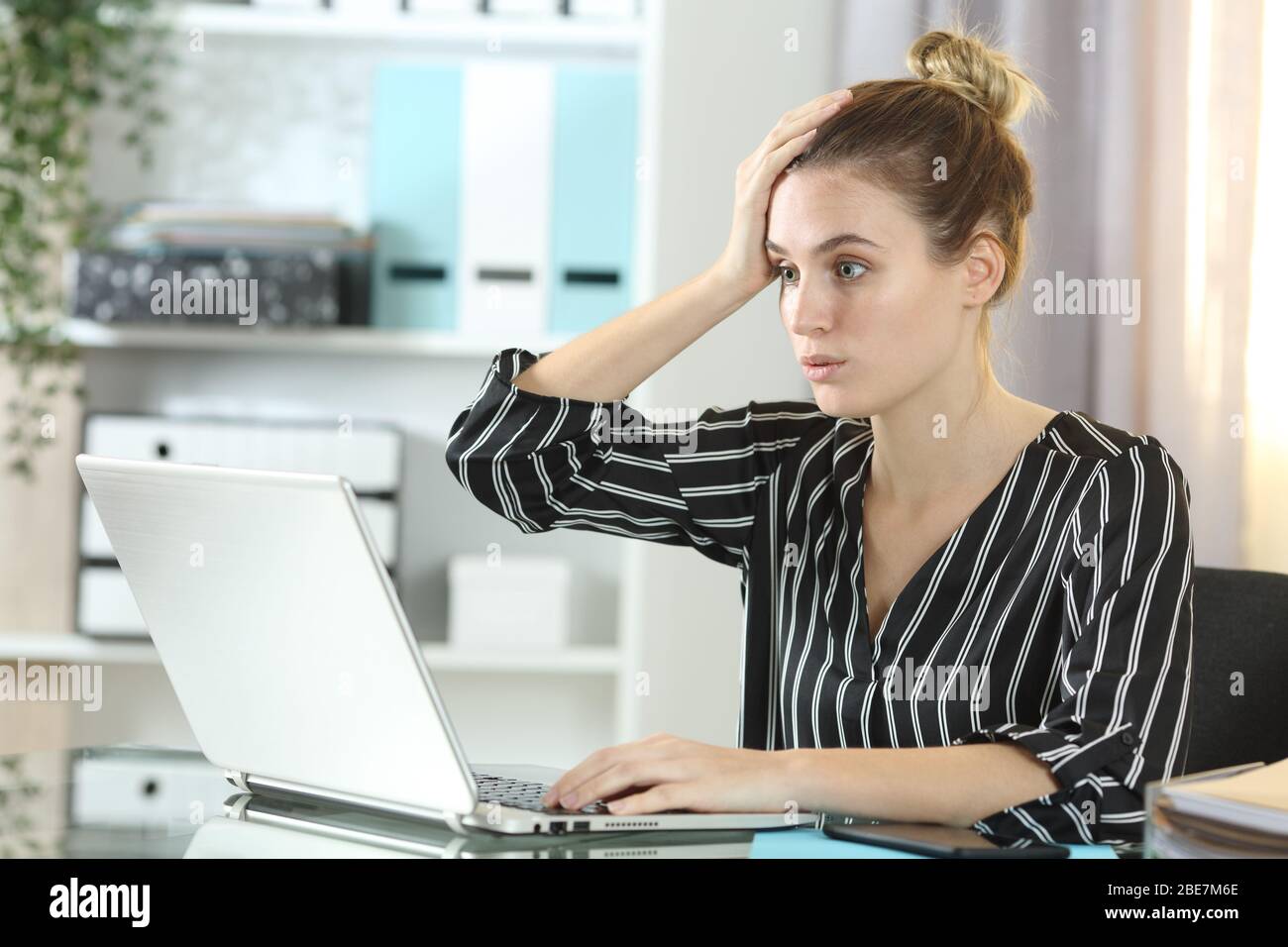 Una donna sorpresa che commette errori online lavorando su un notebook seduto su una scrivania a casa Foto Stock