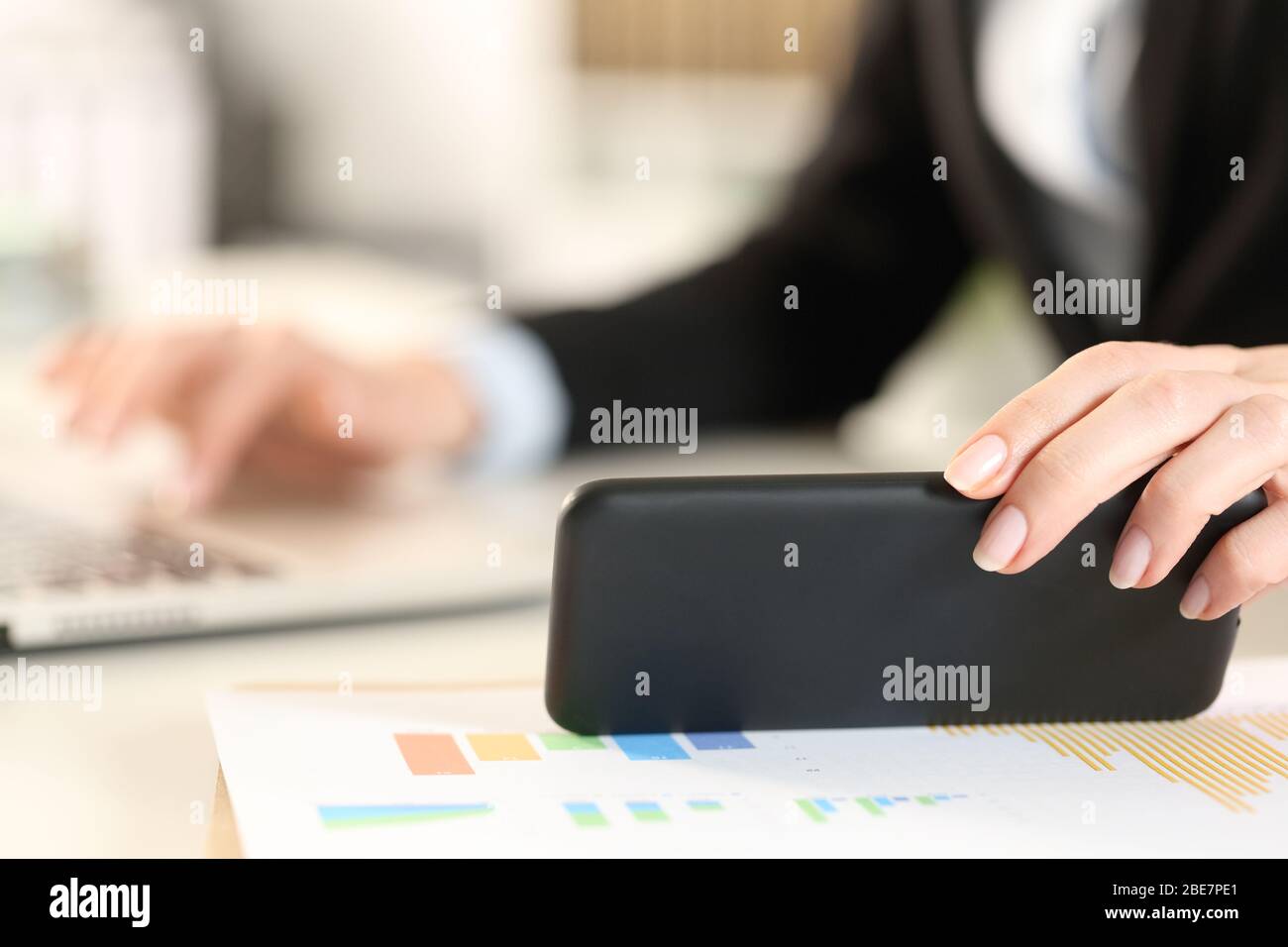Primo piano di una donna executive che utilizza un notebook per controllare la notifica dei messaggi sullo smartphone su una scrivania in ufficio Foto Stock