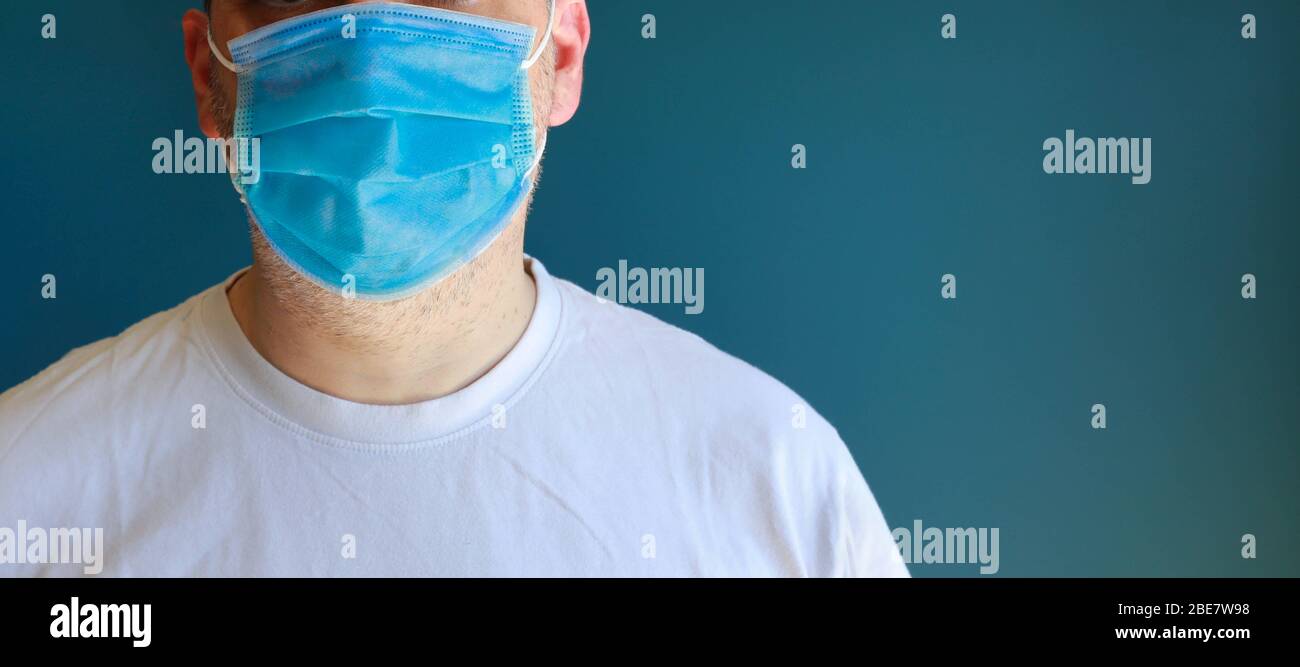 Uomo di mezza faccia che indossa la maschera blu medica durante il blocco epidemico del virus che posa sulla parete blu. Foto Stock