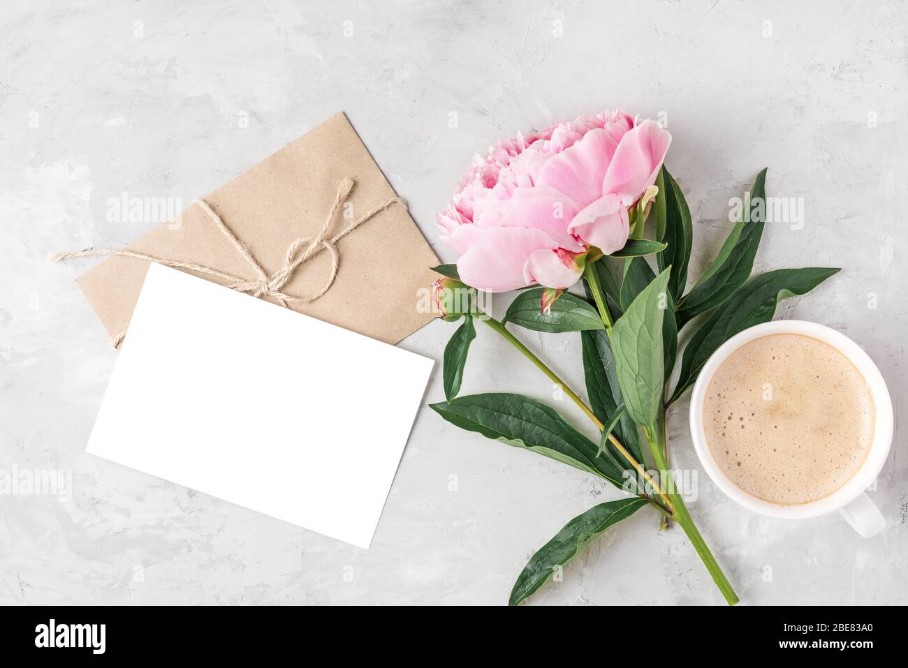 Tazza da caffè del mattino, fiore di peonia rosa e biglietto di auguri vuoto con busta su sfondo chiaro. Mock up. Piatto. Invito al matrimonio. Foto Stock