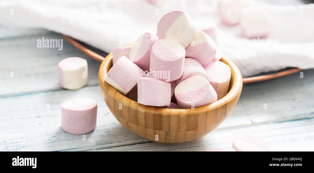 Primo piano di una ciotola di legno piena di marshmallow rosa e bianco con alcuni sparsi intorno su un panno da tavolo bianco, vassoio scuro e tavolo di legno bianco. Foto Stock