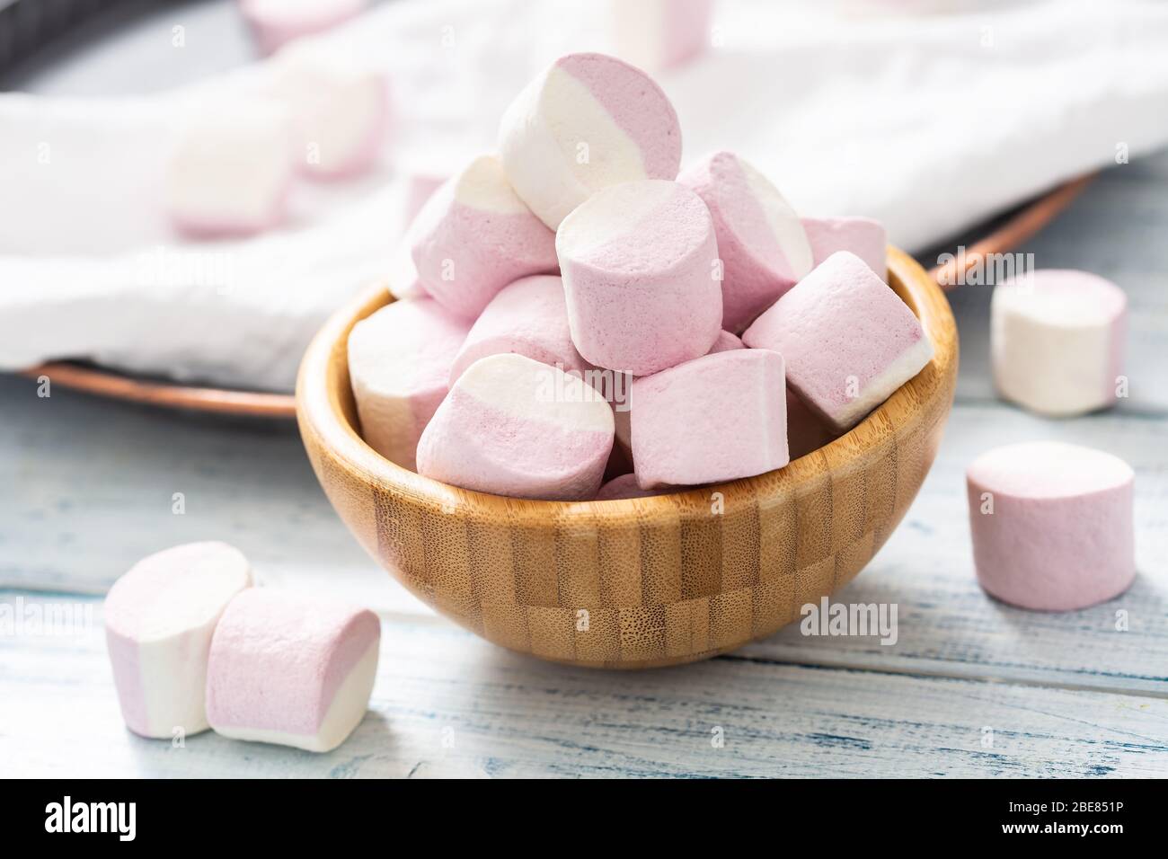 Primo piano di una ciotola di legno piena di marshmallow rosa e bianco con alcuni sparsi intorno su un panno da tavolo bianco, vassoio scuro e tavolo di legno bianco. Foto Stock