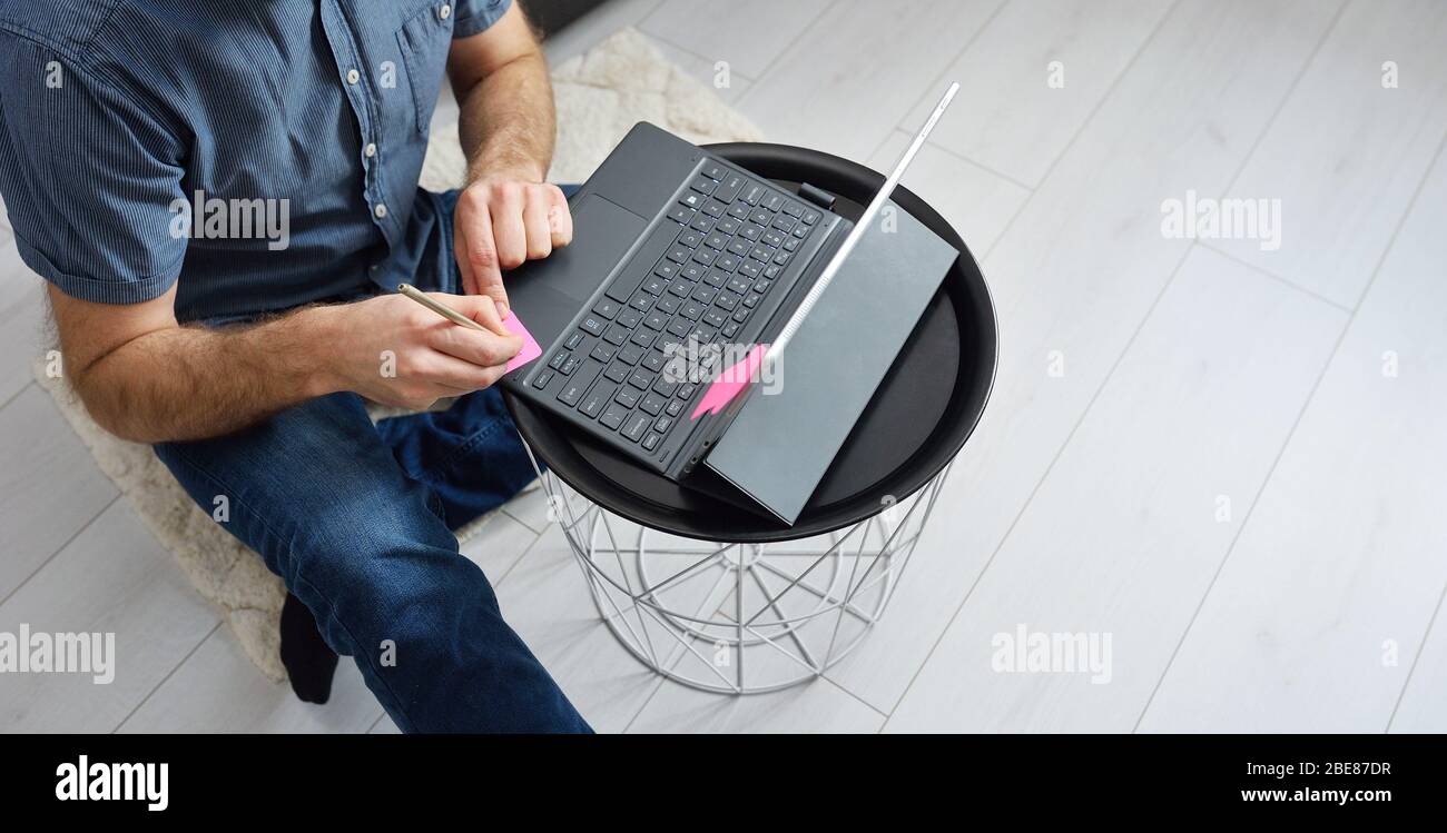 Giovane uomo in abiti casual seduto sul pavimento e lavorando sul suo computer portatile a casa. Rimani a casa e lavora online. Concetto di quarantena, panoramico Foto Stock