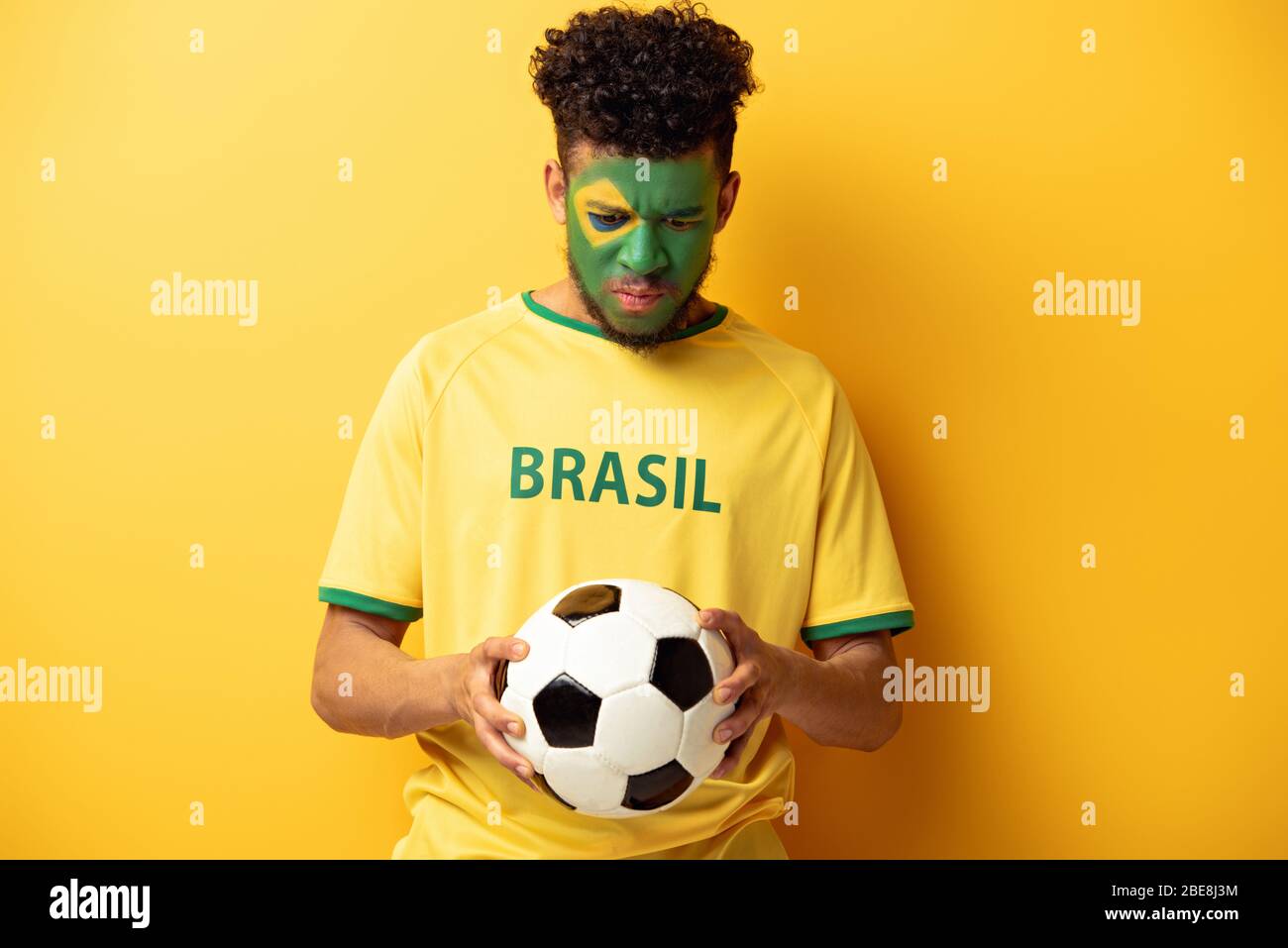 triste fan di football africano americano con faccia dipinta come bandiera brasiliana tenendo palla su giallo Foto Stock
