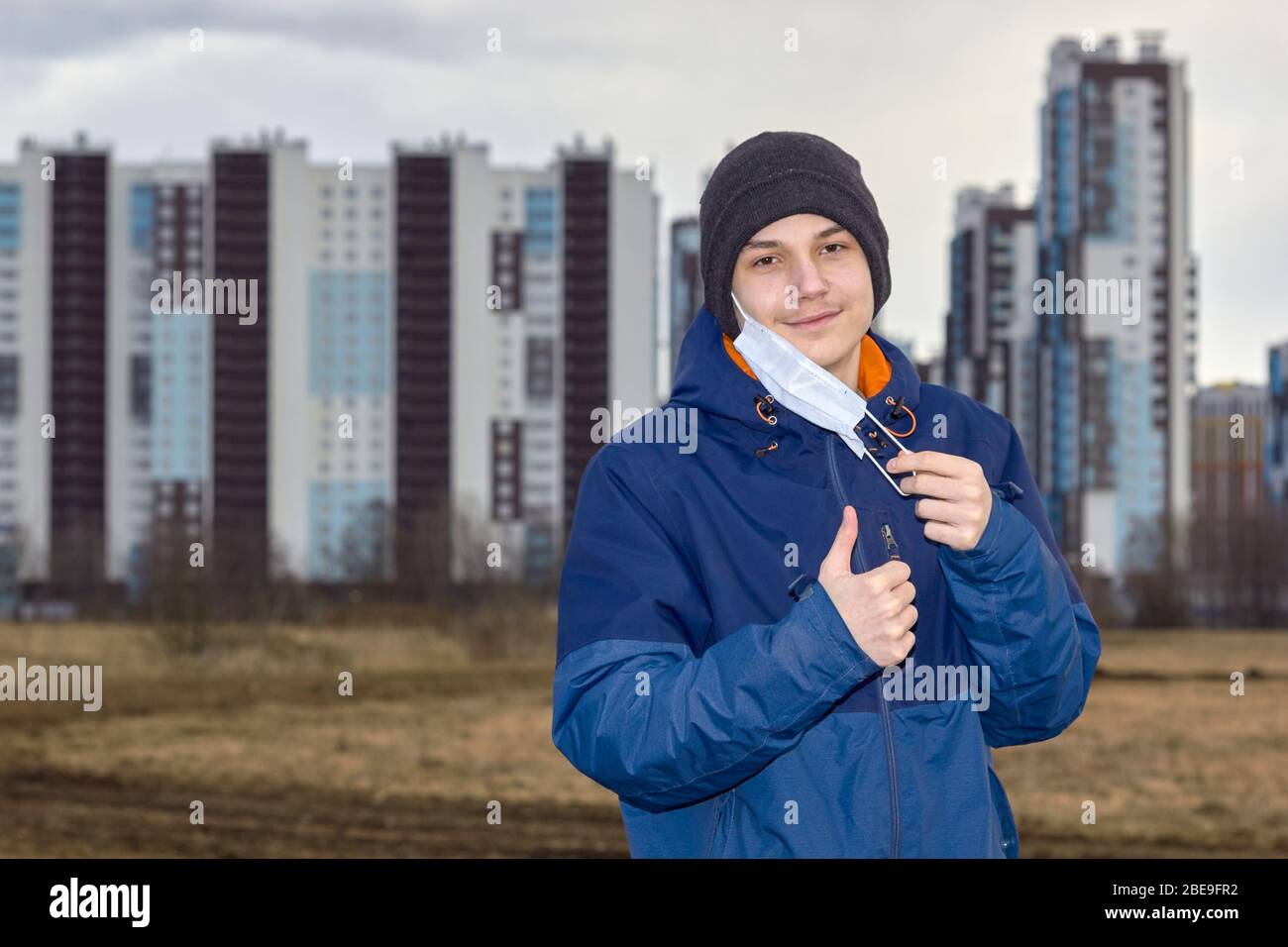 un adolescente in abiti scuri demi-stagione toglie una maschera medica contro lo sfondo di alti blocchi Foto Stock