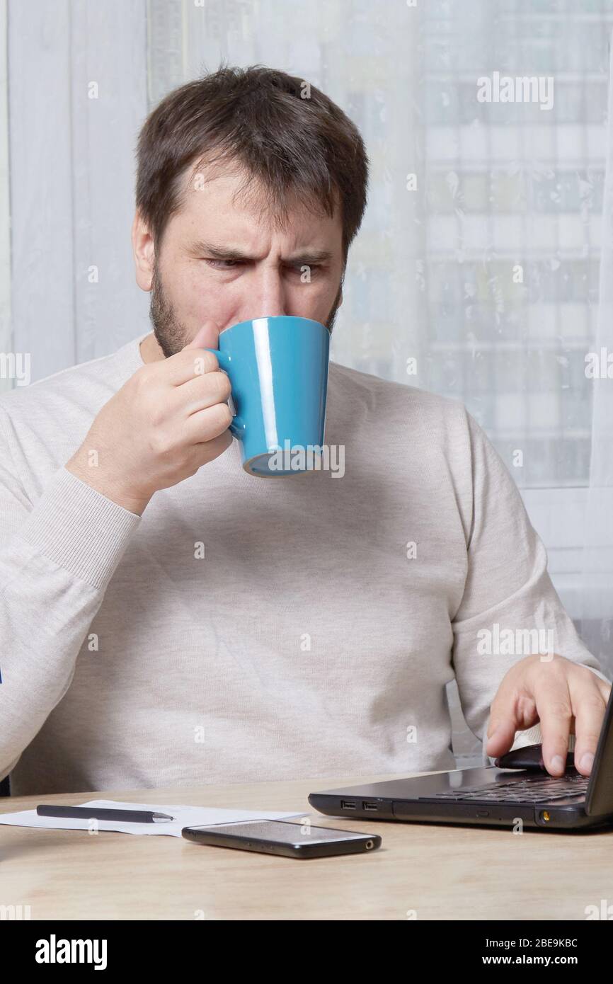 un uomo barbuto è seduto a un tavolo con un computer portatile in un maglione bianco alla ricerca di un lavoro che chiama su un telefono cellulare Foto Stock