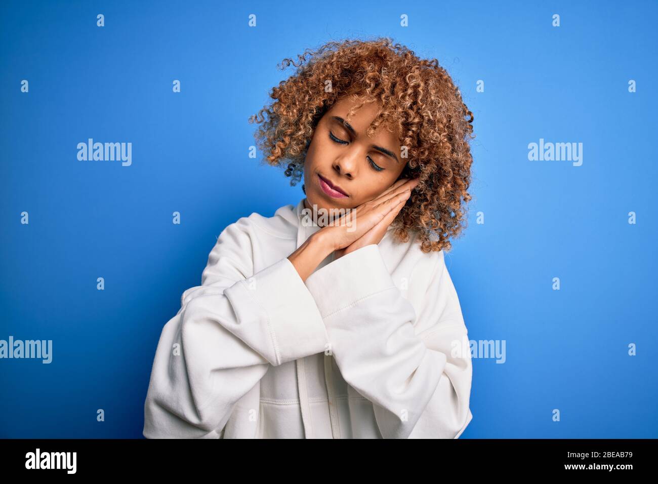Bella donna sportiva african american indossare camicia casual su sfondo blu dormire stanco sognare e posare con le mani insieme mentre SM Foto Stock