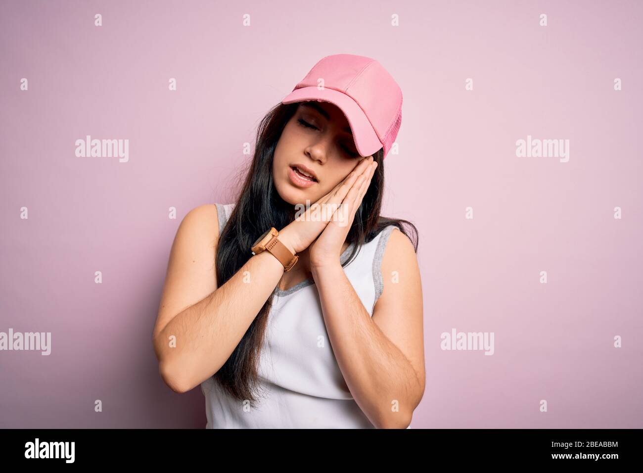 Giovane donna brunetta che indossa cappello sportivo casual su sfondo rosa dormire stanco sognare e posare con le mani insieme mentre sorridente con chiuso ey Foto Stock