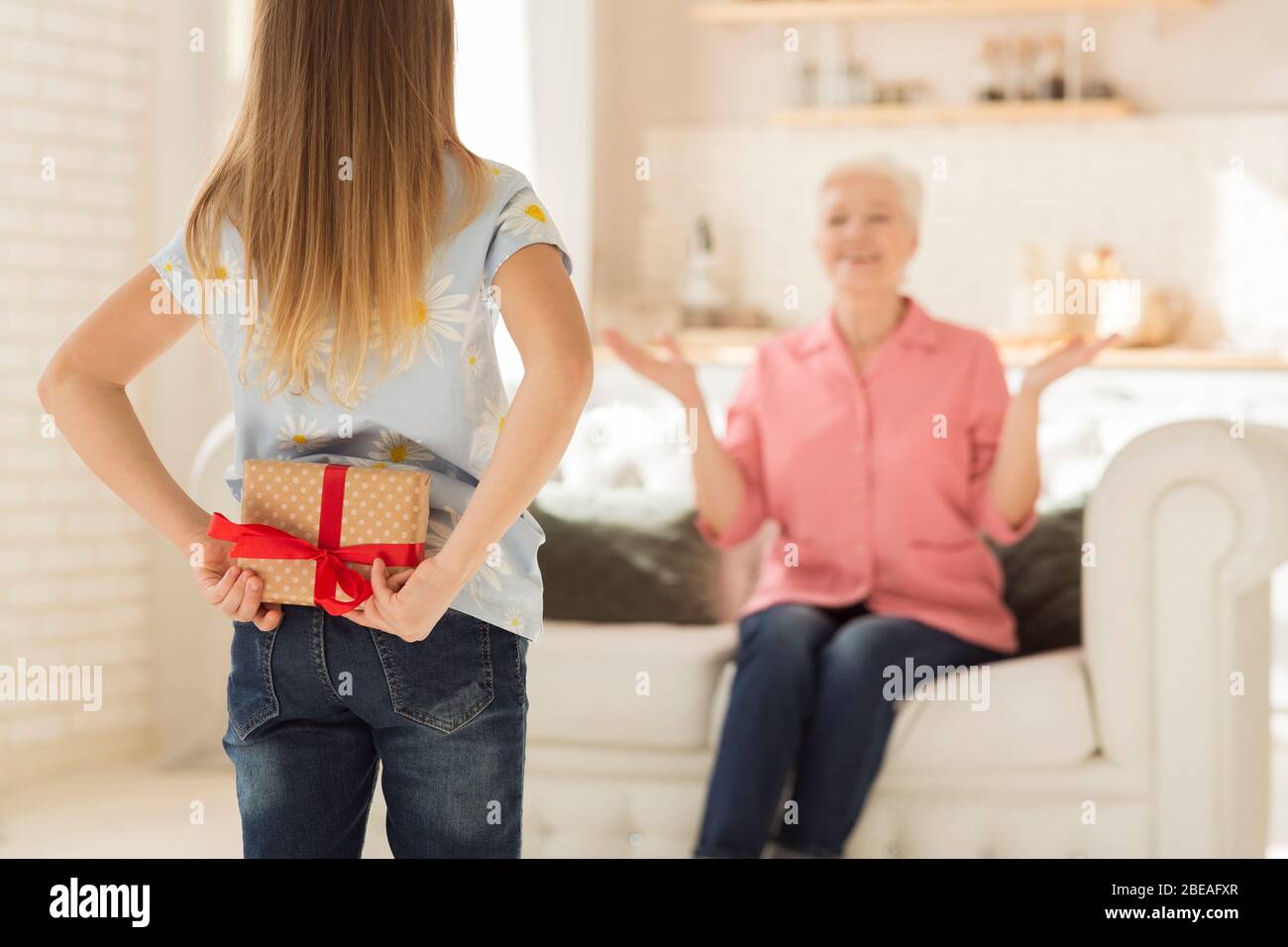 Bambina che nasconde il presente per la nonna dietro la sua schiena, facendo regalo di compleanno a sorpresa a casa. Spazio vuoto Foto Stock