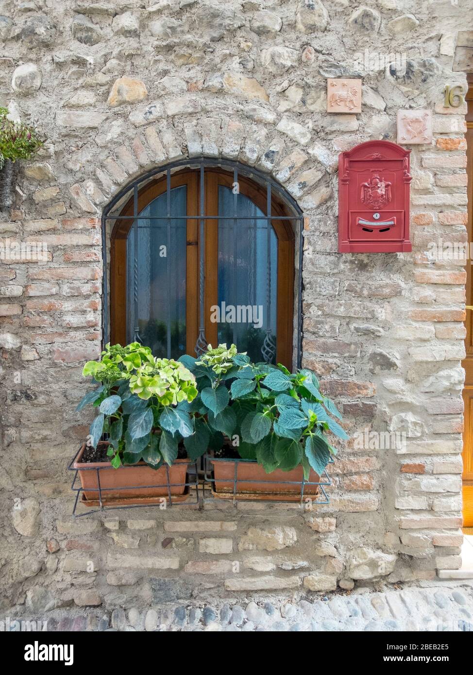 Finestra pittoresca in un complesso storico e restaurato in Italia. Due scatole di fiori con idrangee e una griglia scura. Una casella di posta rossa cattura l'occhio. Foto Stock