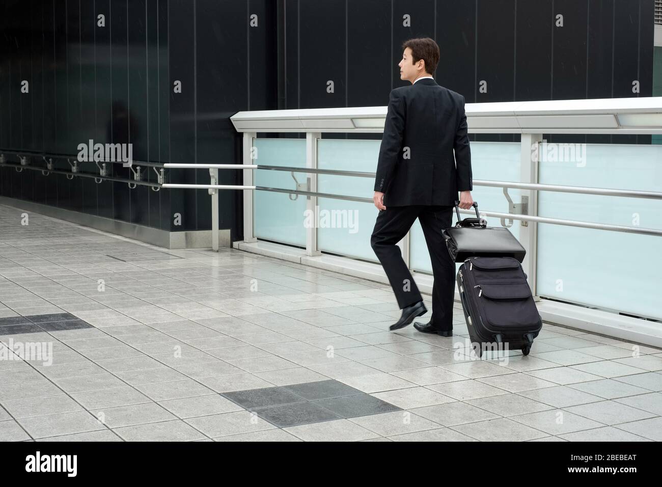 Un uomo d'affari vestito in un vestito nero con una valigia a rotelle (a mano) e una valigetta. Foto Stock