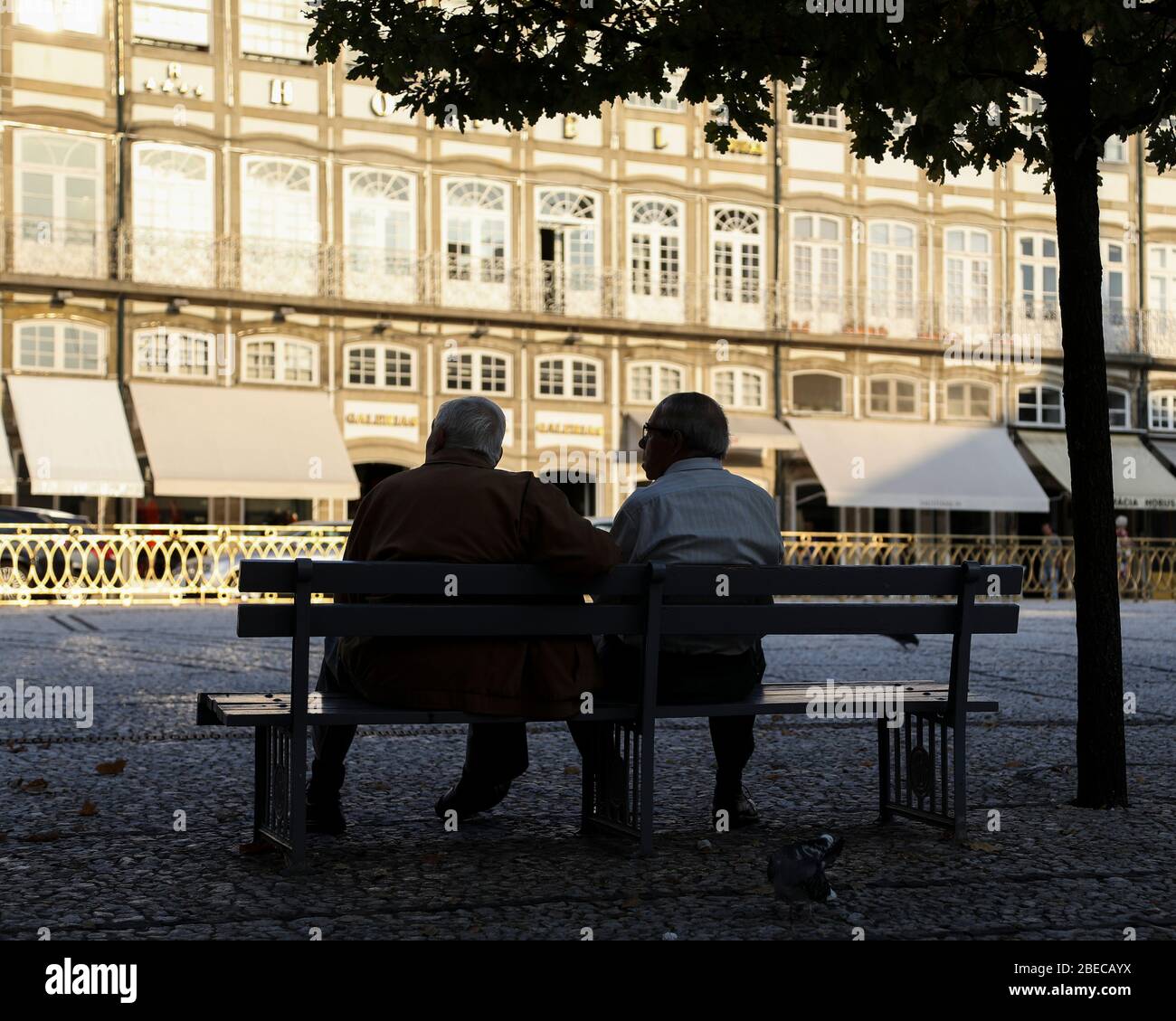 Due vecchi signori seduti su una panchina che guardano il mondo passare nella piazza della città. Foto Stock
