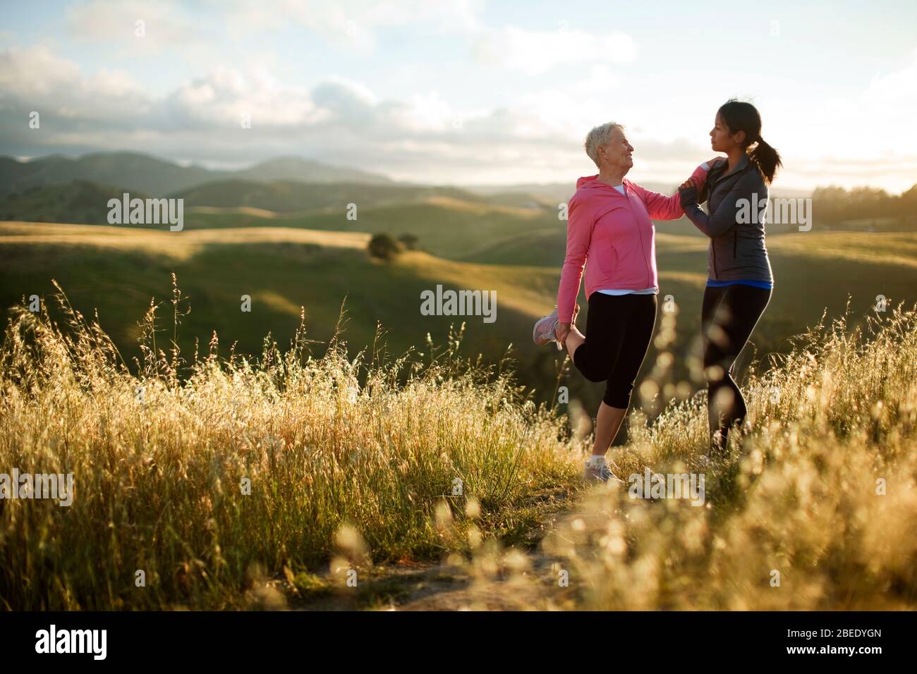 Una giovane donna che aiuta una donna anziana a allungarsi mentre si esercita in terreno collinare Foto Stock