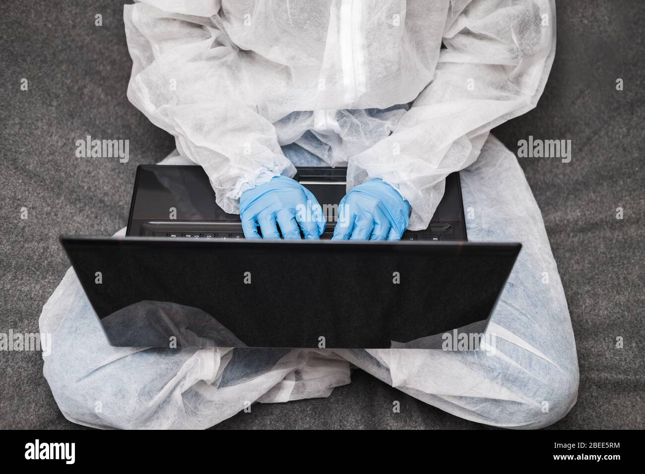 Persona in tuta protettiva bianca traslucida, guanti in gomma blu, maschera medica si siede al tavolo a casa e lavora o studia sul laptop durante la quarantena. Foto Stock