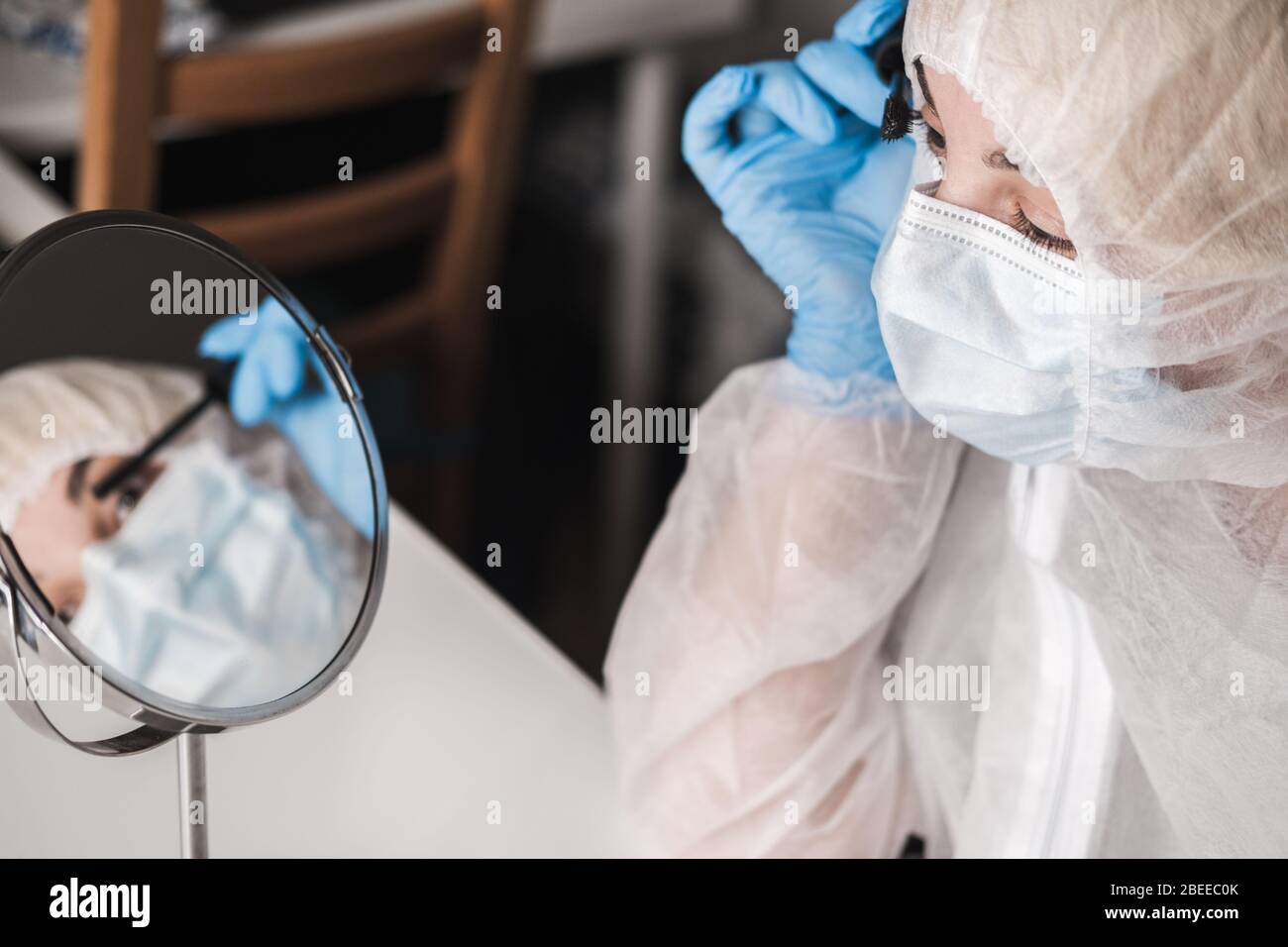 Ragazza in tuta trasparente bianca protettiva, guanti di gomma blu, maschera medica si siede a casa del tavolo e dipinge ciglia con mascara allo specchio durante Foto Stock