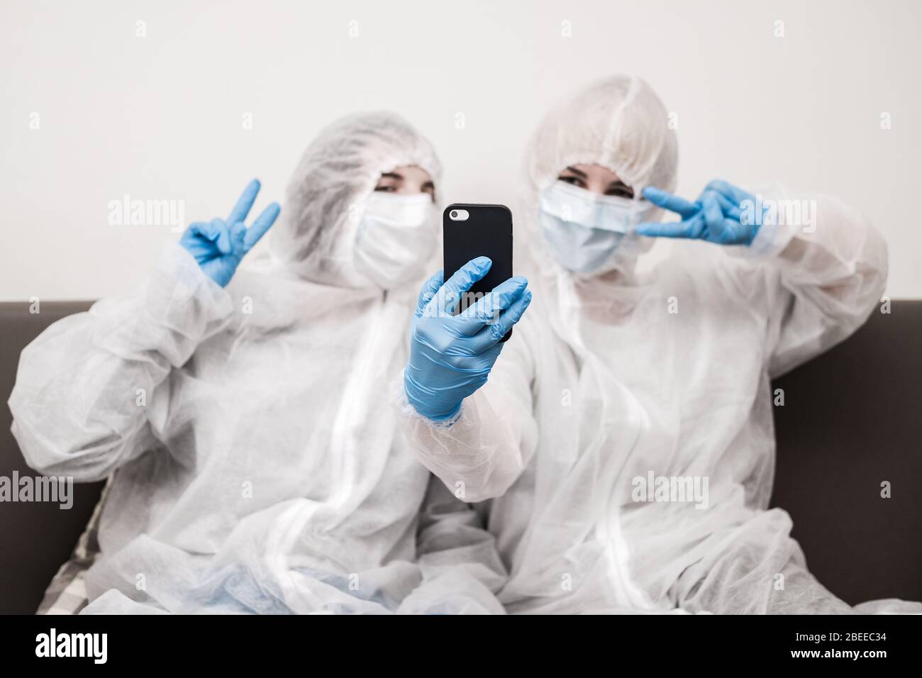 Due persone in tuta protettiva bianca traslucida, guanti in gomma blu e maschera medica sono seduti sul divano a casa e con il selfie sullo smartphone. Rimani a casa Foto Stock