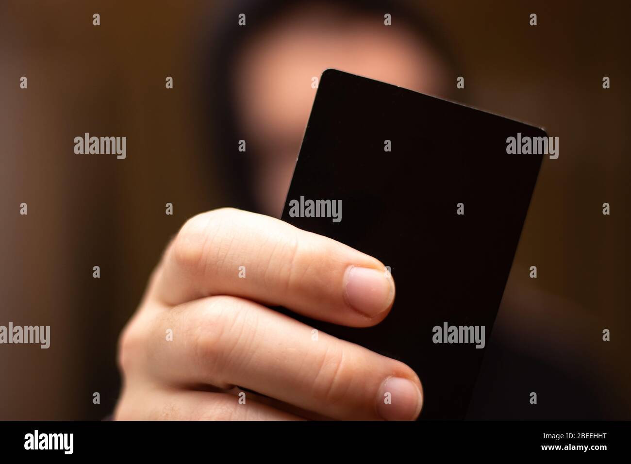 Scura strana immagine di un uomo sfocato sullo sfondo in una felpa con cappuccio che tiene una carta nera vuota nelle sue dita Foto Stock