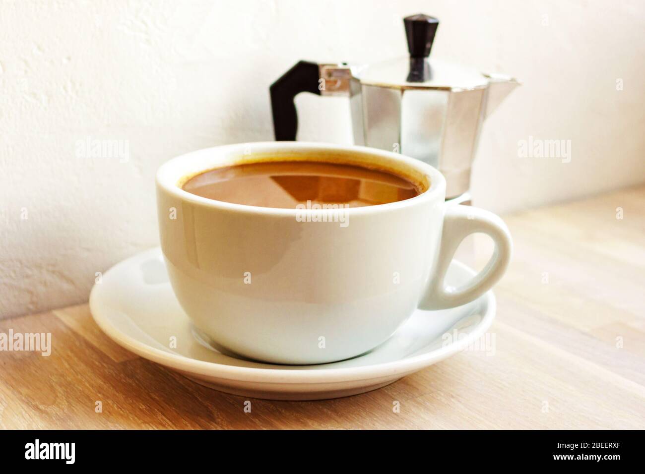 Tazza di caffè al latte con piattino, macchina da caffè in alluminio su sfondo di legno chiaro. Colazione. Foto Stock