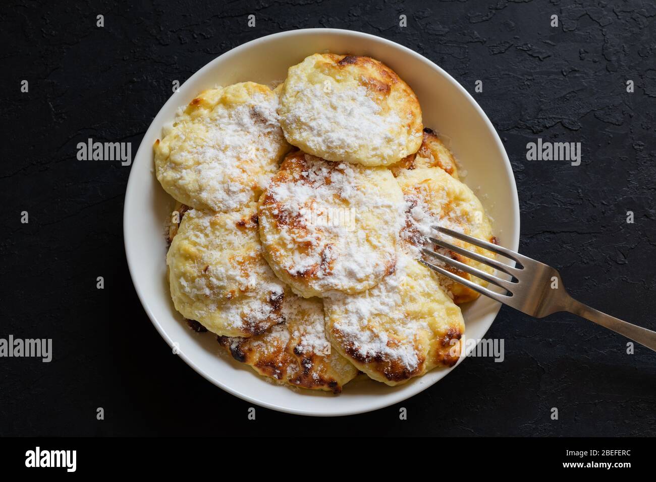 Dolci pancake di formaggio casolare su un piatto bianco cosparso di zucchero in polvere con una forchetta su sfondo scuro. Facile da preparare la colazione fatta in casa. Foto Stock