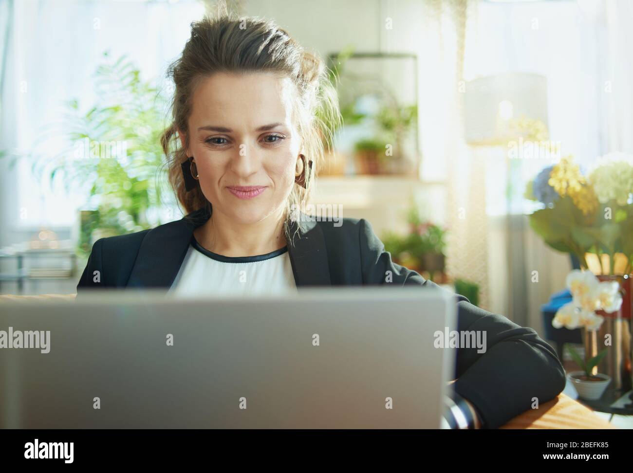 elegante casalinga di mezza età in blusa bianca e giacca nera nella casa moderna in giorno di sole surf web su un computer portatile mentre si siede sul divano. Foto Stock