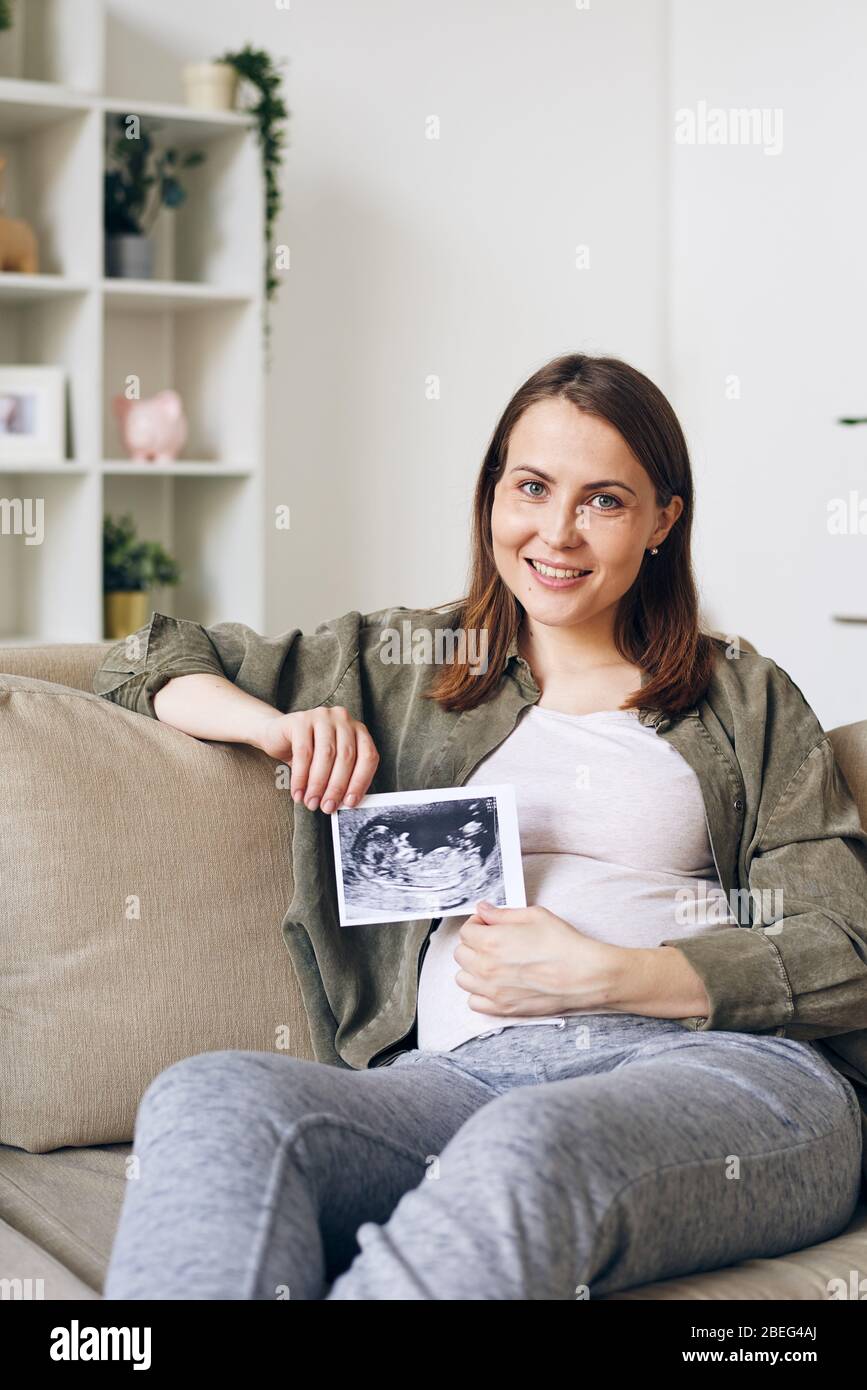 Ritratto di sorridente bella giovane donna incinta seduta in posa rilassata su divano e posa con foto ecografica del bambino Foto Stock