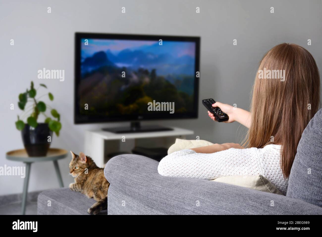 Una giovane donna che guarda la TV mentre si siede con il suo gatto sul divano nel soggiorno a casa. Natura, documentario, schermo tv, binge watching Foto Stock