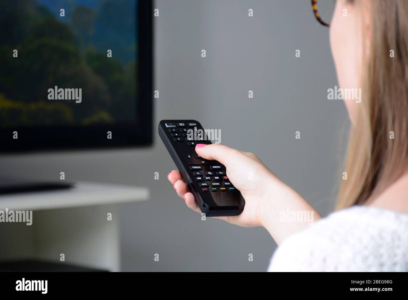Una giovane donna con occhiali che guardano la TV e cambia canale con telecomando tv. Natura, documentario, schermo tv, binge watching Foto Stock