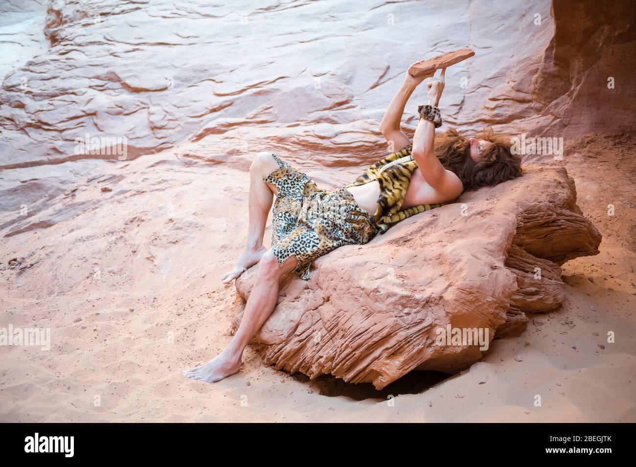 Caveman si distende da solo utilizzando la sua primitiva tavoletta di pietra all'aperto in una grotta di roccia intemperata Foto Stock