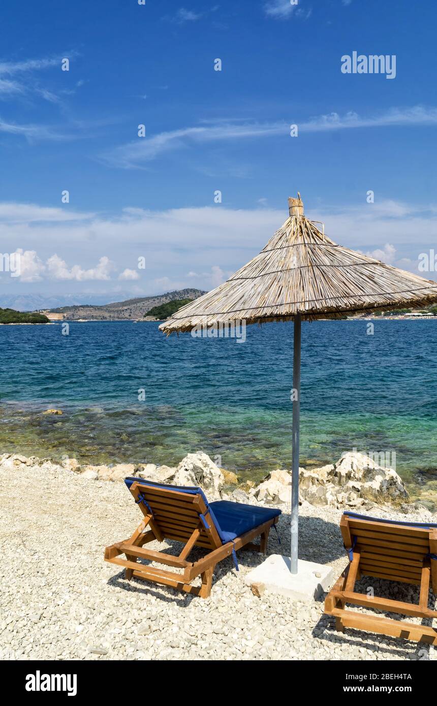 Spiaggia rocciosa sulla riva del Mar Ionio con lettini da mare e ombrelloni di paglia. Il blu e il mare tourquse in background, nessuno. Soleggiata giornata estiva. Ksamil, Al Foto Stock