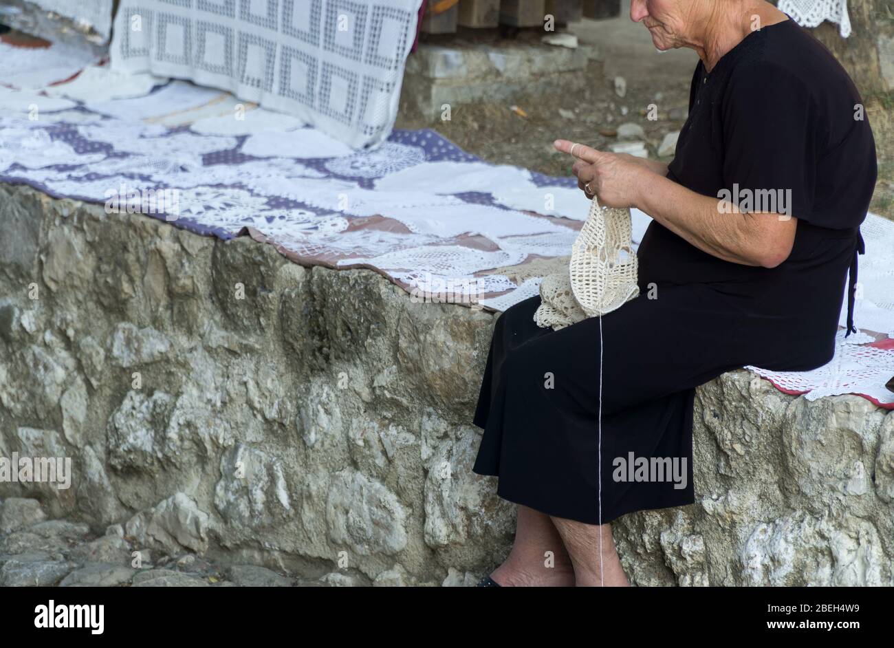 Una vecchia donna che indossa un abito nero rende tovaglioli di pizzo decorativo con un crochet. Lavorazione dell'ago, accovacciamento. KRUJE, ALBANIA, 19 SETTEMBRE 2019. Foto Stock